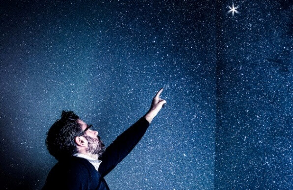 Un hombre, en una habitación empapelada, como si fuera el firmamento por la noche, señalando una estrella que brilla más que el resto.